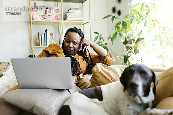 Frau liegt auf dem Sofa mit Hund und arbeitet am Laptop