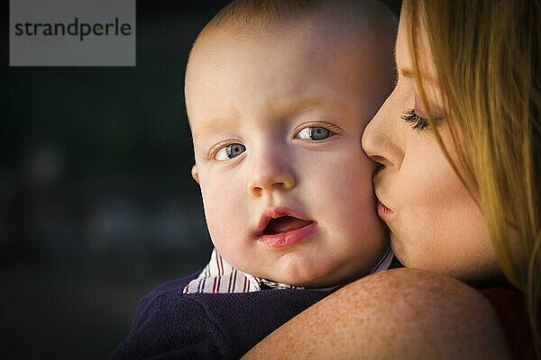 Adorable roten Kopf Säugling Junge wird von seiner Mutter im Freien in dramatischen Beleuchtung geküsst