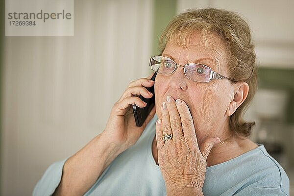 Geschockte ältere Frau am Handy mit Hand über dem Mund in der Küche