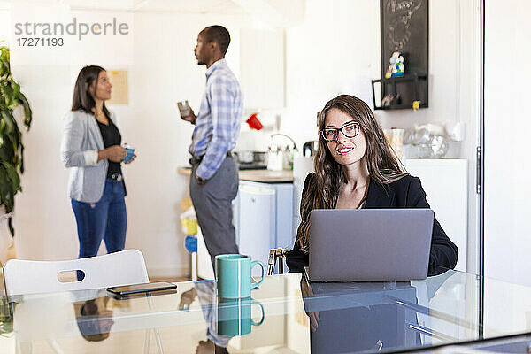 Lächelnde Geschäftsfrau  die mit einem Laptop am Tisch sitzt  während im Hintergrund Kollegen in der Kaffeepause diskutieren