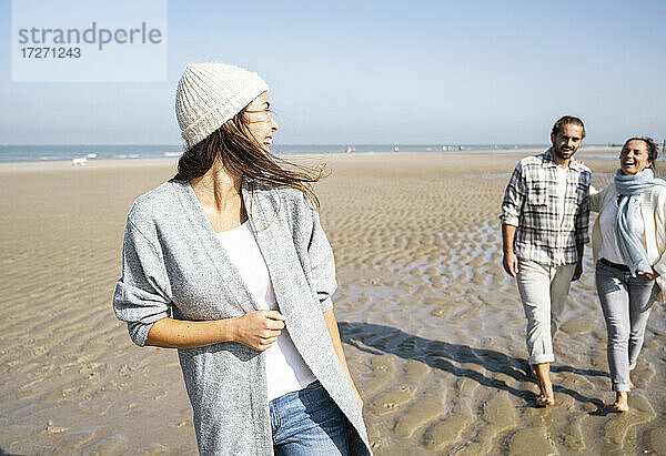 Junge Frau lacht und sieht einen Mann und eine Mutter  die im Hintergrund am Strand spazieren gehen