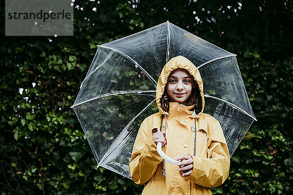 Mädchen mit Regenmantel  das einen Regenschirm hält  während es an einer Laubwand steht