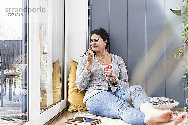 Lächelnde junge Frau isst Erdbeere  während sie zu Hause am Fenster sitzt