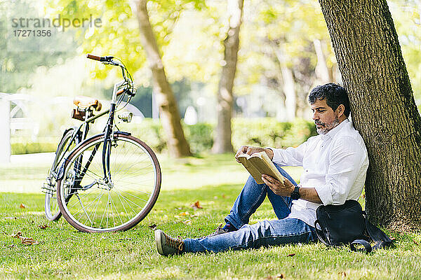 Älterer Mann liest ein Buch und lehnt sich an einen Baumstamm in einem öffentlichen Park