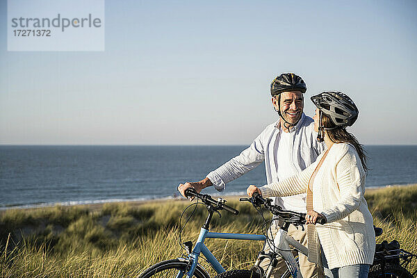 Glückliches Paar in Fahrradhelmen steht mit Fahrrad am Strand gegen klaren Himmel