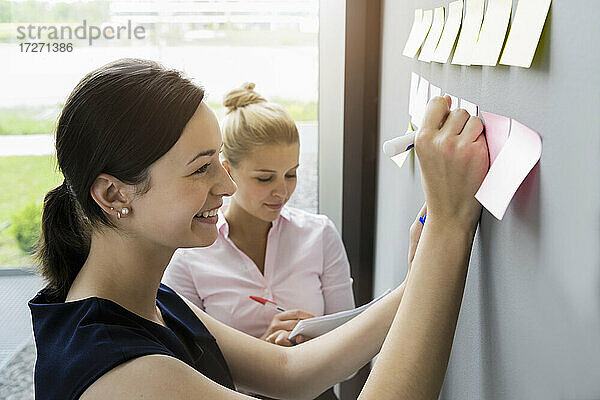 Lächelnde Geschäftsfrau  die auf einen an die Wand geklebten Klebezettel schreibt  während sie mit einem Kollegen im Büro diskutiert