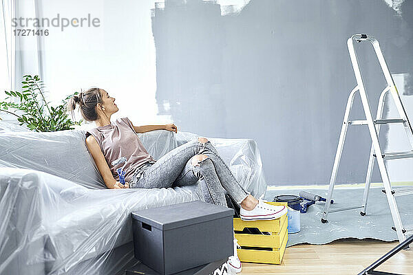 Frau mit Farbrolle betrachtet die Wand  während sie zu Hause auf dem Sofa sitzt