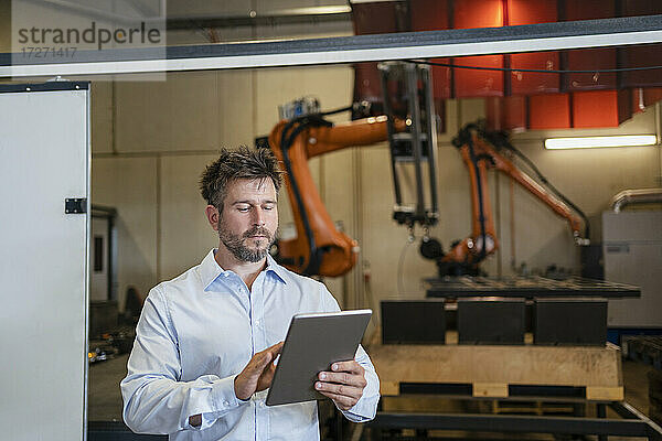 Reifer Geschäftsmann  der ein digitales Tablet benutzt  während er in einer Fabrik vor einer Roboterarm-Maschine steht