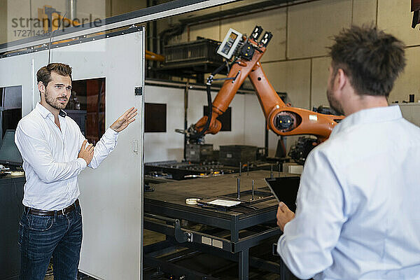 Geschäftsmann zeigt auf eine Roboterarm-Maschine  während er mit einem Kollegen in einer Fabrik steht