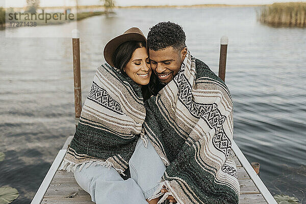Lächelndes  in eine Decke gehülltes Paar sitzt auf einem Steg am See