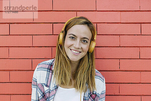Lächelnde Frau  die über einen Kopfhörer Musik hört  an einer Backsteinmauer