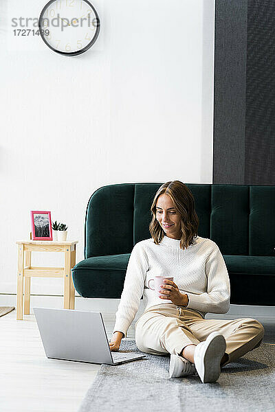 Geschäftsfrau mit Kaffeetasse und Laptop  während sie im Büro auf dem Boden sitzt