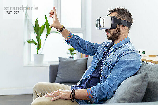 Mittlerer erwachsener Mann  der einen Virtual-Reality-Simulator trägt und auf dem Sofa sitzt