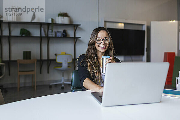 Lächelnde Geschäftsfrau  die Kaffee trinkt und einen Laptop am Schreibtisch im Büro benutzt