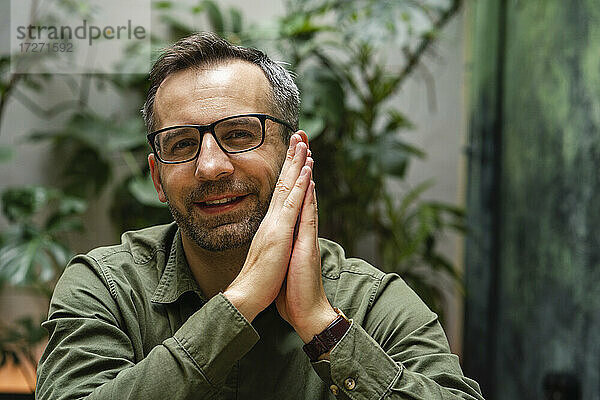 Lächelnder Geschäftsmann mit Brille sitzt mit verschränkten Händen im Cafe