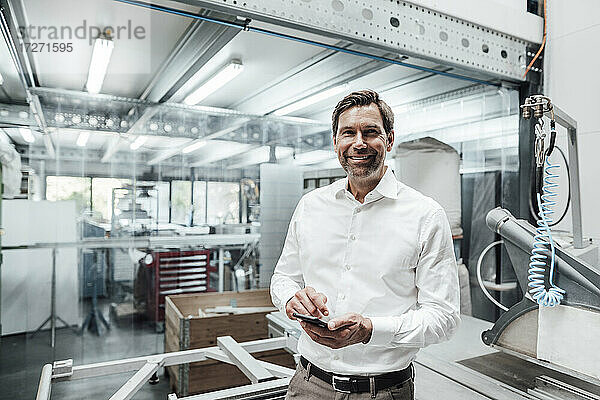 Lächelnder männlicher Ingenieur mit Smartphone an einer Maschine in der Industrie