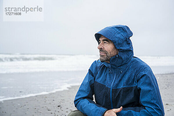 Nachdenklicher reifer Mann in blauem Regenmantel  der am Strand vor dem Himmel kauert