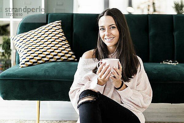 Lächelnde Frau sitzt mit Kaffeetasse auf dem Sofa zu Hause