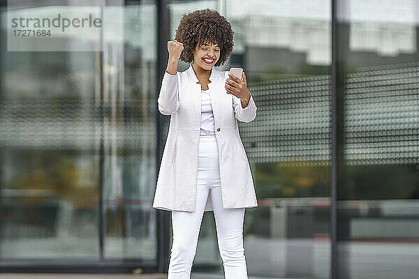 Junge Geschäftsfrau  die eine gewinnende Geste zeigt  während sie ein Mobiltelefon benutzt und im Freien steht