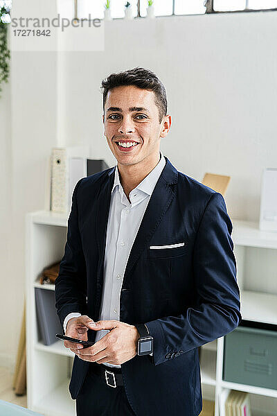 Lächelnder  gutaussehender Geschäftsmann  der ein Smartphone hält  während er an einem kreativen Arbeitsplatz steht