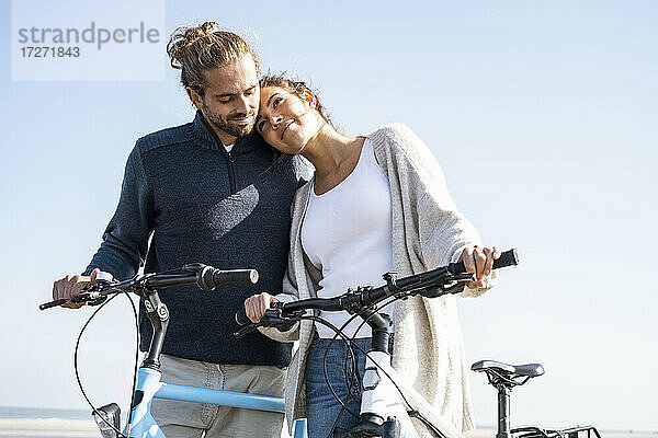 Schöne Frau mit Kopf auf der Schulter ihres Freundes steht mit Fahrrädern am Strand gegen klaren Himmel an einem sonnigen Tag