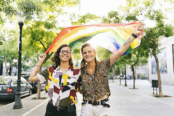 Glückliches lesbisches Paar schwenkt Regenbogenfahne auf dem Fußweg in der Stadt