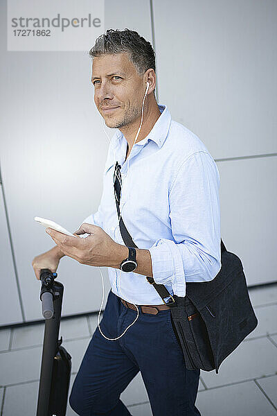 Älterer Geschäftsmann  der ein Smartphone benutzt und über Kopfhörer Musik hört  während er auf einem Elektroroller steht