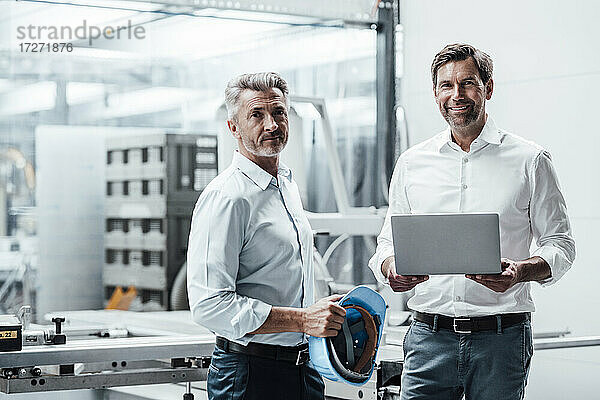 Lächelnder männlicher Ingenieur mit Laptop  der neben einem Kollegen mit Schutzhelm in einer Fabrik steht