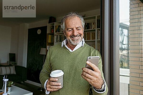 Lächelnder reifer Mann mit Kaffeetasse  der ein Mobiltelefon benutzt  während er zu Hause steht