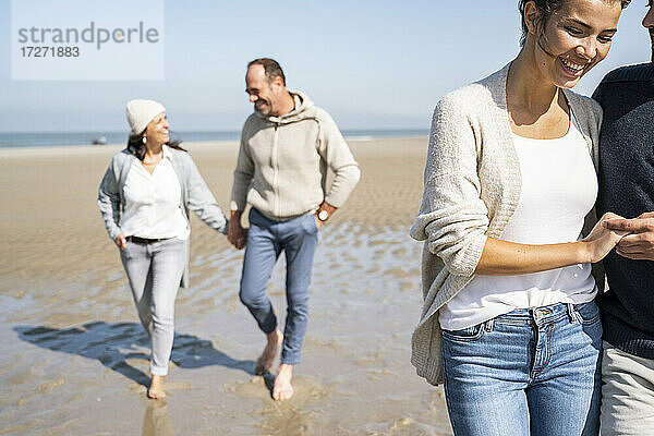 Lächelnde Frau  die einen Mann an der Hand hält  während sie mit einem Paar im Hintergrund am Strand spazieren geht