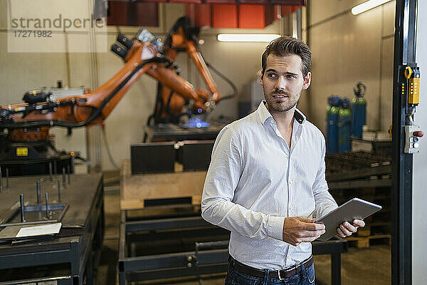 Junger Geschäftsmann  der ein digitales Tablet benutzt  während er vor einer Roboterarm-Maschine in einer modernen Fabrik steht