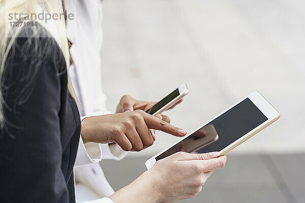 Geschäftsfrau  die auf ein digitales Tablet zeigt  während sie das Mobiltelefon eines Kollegen in der Stadt benutzt