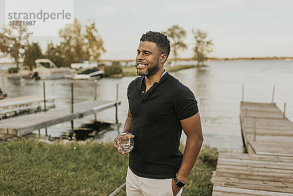 Lächelnder Mann mit Weinglas und Hand in der Tasche an der Seebrücke stehend