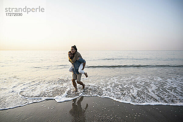 Fröhlicher Mann  der seine Freundin huckepack nimmt  während er am Strand gegen den klaren Himmel bei Sonnenuntergang spazieren geht