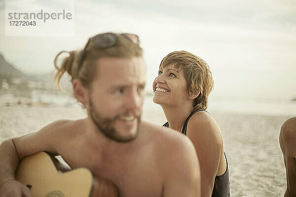Lächelnder Mann spielt Gitarre am Strand sitzend an einem sonnigen Tag