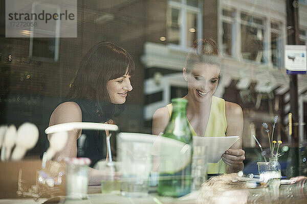 Lächelnde Geschäftsfrauen  die während eines Treffens in einem Café über ein Dokument diskutieren  gesehen durch ein Fensterglas