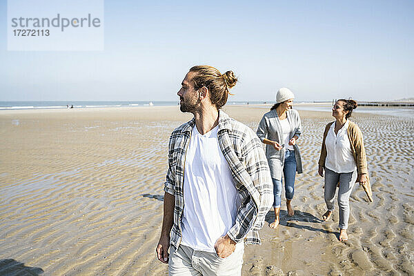 Mann schaut weg  während er mit Frauen im Hintergrund am Strand spazieren geht