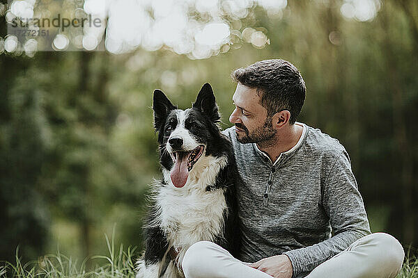 Lächelnder Mann  der seinen Hund anstarrt  während er in einem öffentlichen Park sitzt