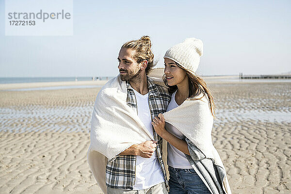 Nachdenkliches junges Paar  das in eine Decke gehüllt ist und wegschaut  während es am Strand an einem sonnigen Tag steht