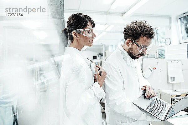 Eine Wissenschaftlerin steht neben einem männlichen Kollegen  der einen Laptop im Labor benutzt