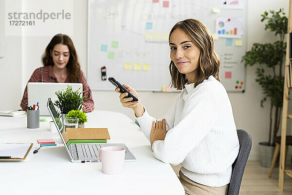 Lächelnde Geschäftsfrau  die ein Mobiltelefon benutzt  während ihr Kollege im Hintergrund an einem Laptop arbeitet