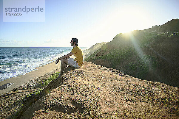 Mann sitzt auf einer Felsformation und betrachtet das Meer vor dem Himmel