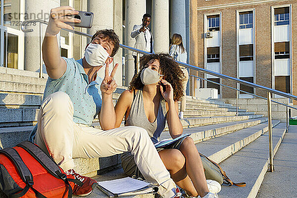 Männlicher Freund gestikuliert Frieden  während er ein Selfie mit einem Universitätsstudenten mit Schutzmaske auf einer Treppe in der Stadt macht