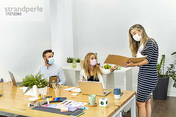 Angestellter mit Gesichtsmaske bei der Besprechung einer Akte während der Arbeit mit einem Kollegen im Büro
