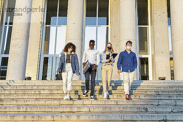 Universitätsstudenten tragen einen Gesichtsschutz  während sie die Treppe vor dem Bildungsgebäude hinuntergehen