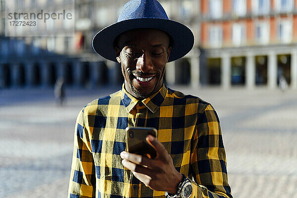 Lächelnder afrikanischer Mann mit Hut  der in der Stadt ein Smartphone benutzt