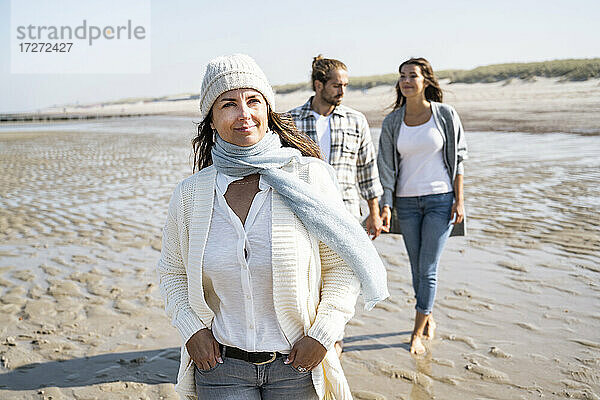 Lächelnde Frau  die wegschaut  während sie mit einem Paar am Strand spazieren geht  das sich an den Händen hält  im Hintergrund