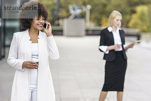 Geschäftsfrau  die mit einem Mobiltelefon spricht  während sie mit einem Kollegen im Hintergrund auf dem Fußweg steht