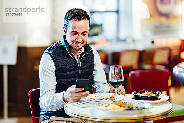 Lächelnder junger Mann  der im Restaurant sitzt und mit seinem Smartphone SMS schreibt