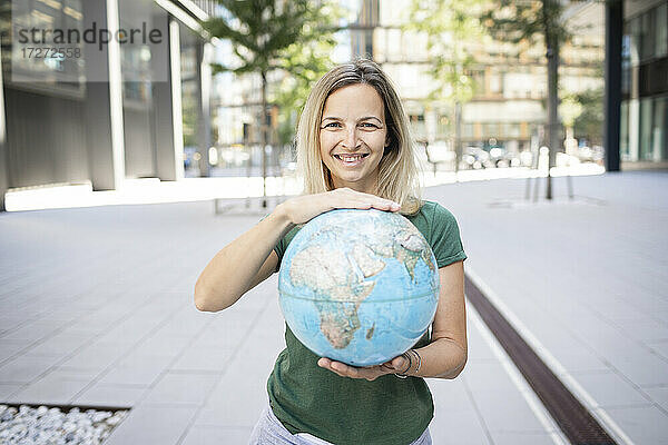 Lächelnde Frau  die einen Globus hält  während sie auf einem Fußweg in der Stadt steht